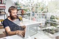 Un bărbat cu barbă stă la un birou și folosește un laptop în contextul serviciilor IT ale CANCOM Austria.