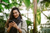 Femeie în aer liber zâmbind la un telefon mobil, înconjurată de copaci.