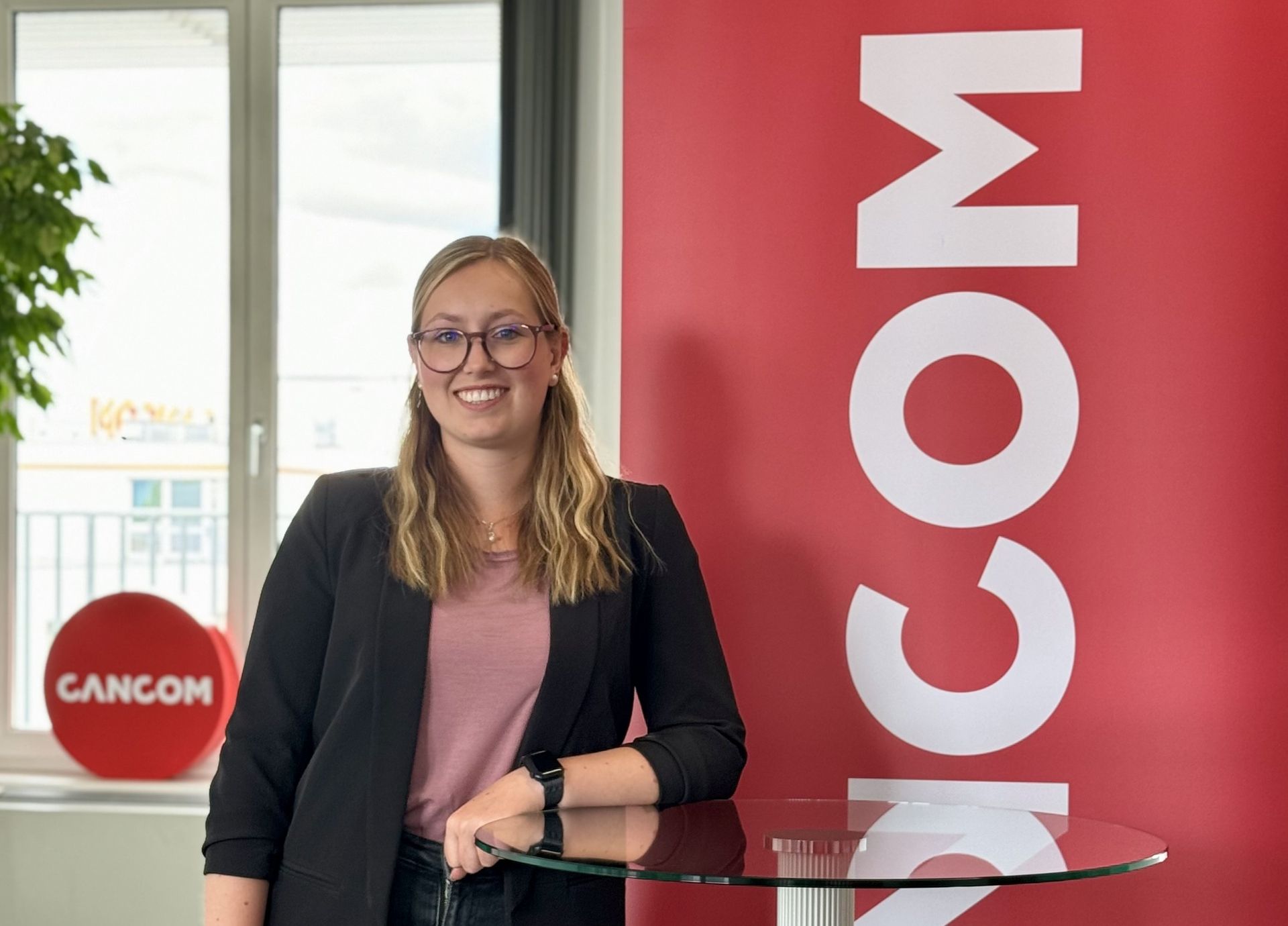 Femeie cu ochelari zâmbește în picioare în fața unui semn.