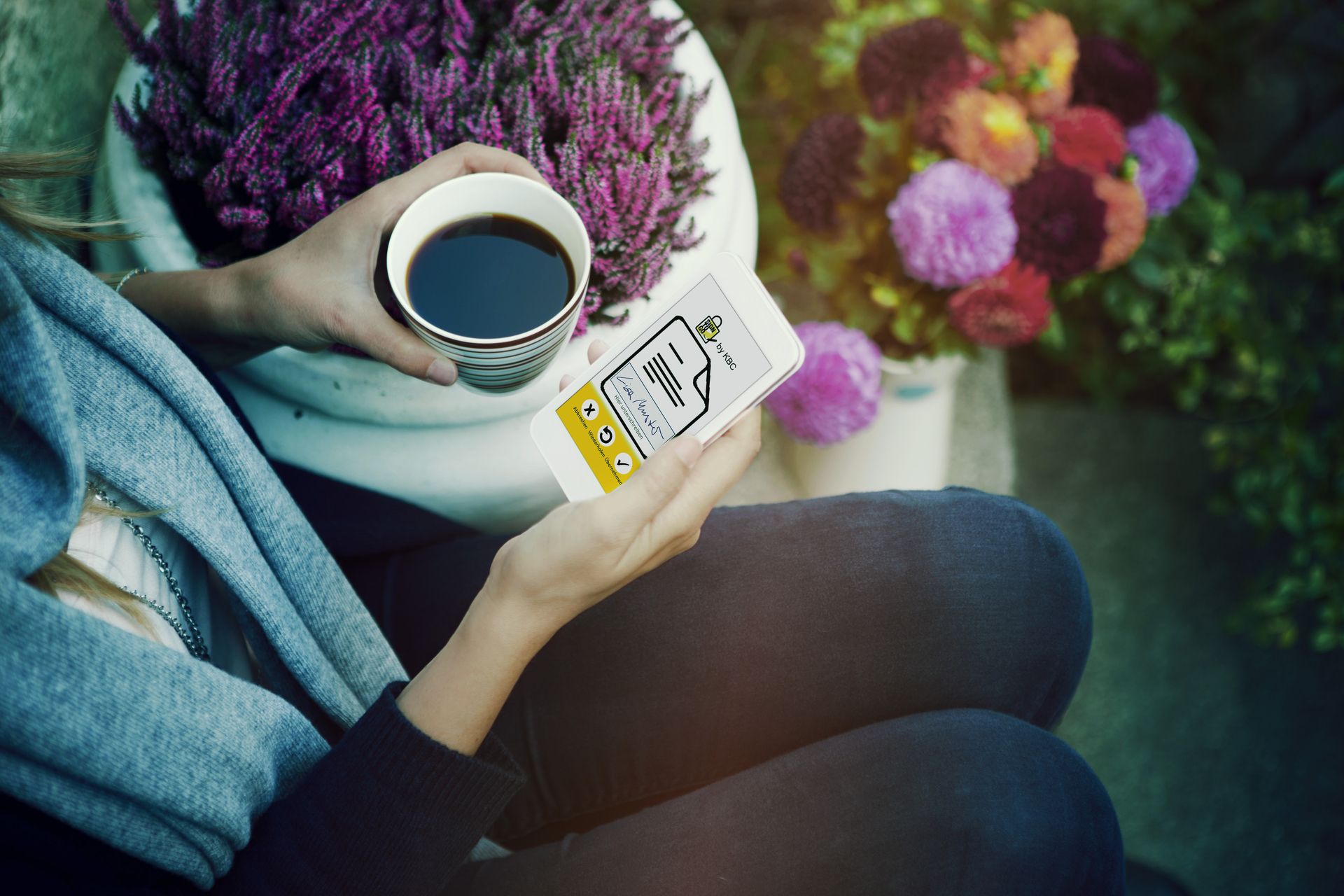 Persoană care ține în mână o ceașcă de cafea și un smartphone, înconjurată de flori în aer liber.