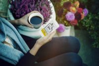 Persoană care ține în mână o ceașcă de cafea și un smartphone, înconjurată de flori în aer liber.