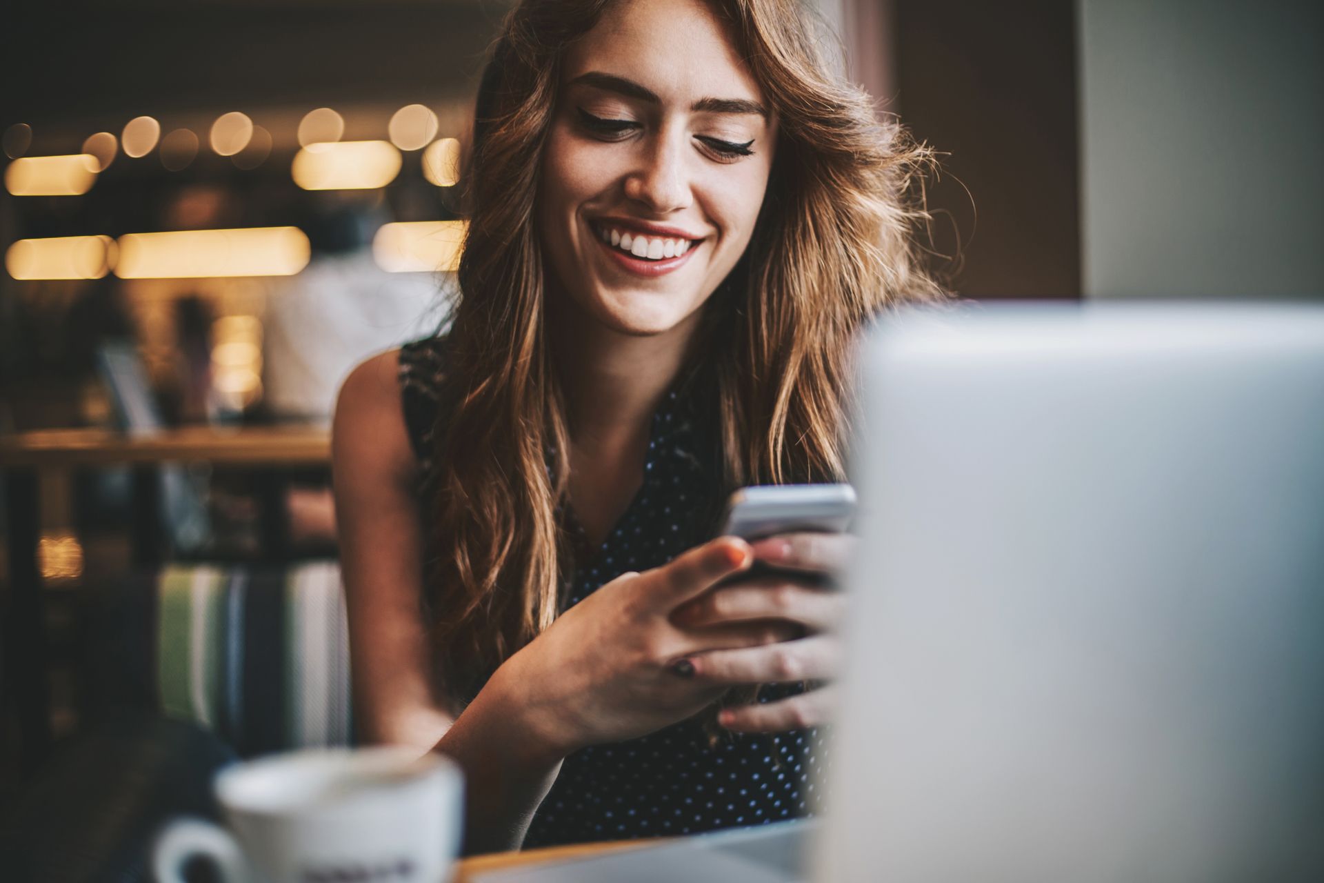 Femeie zâmbește în timp ce se uită la smartphone-ul ei într-o configurație modernă de birou acasă cu laptop și ceașcă de cafea.